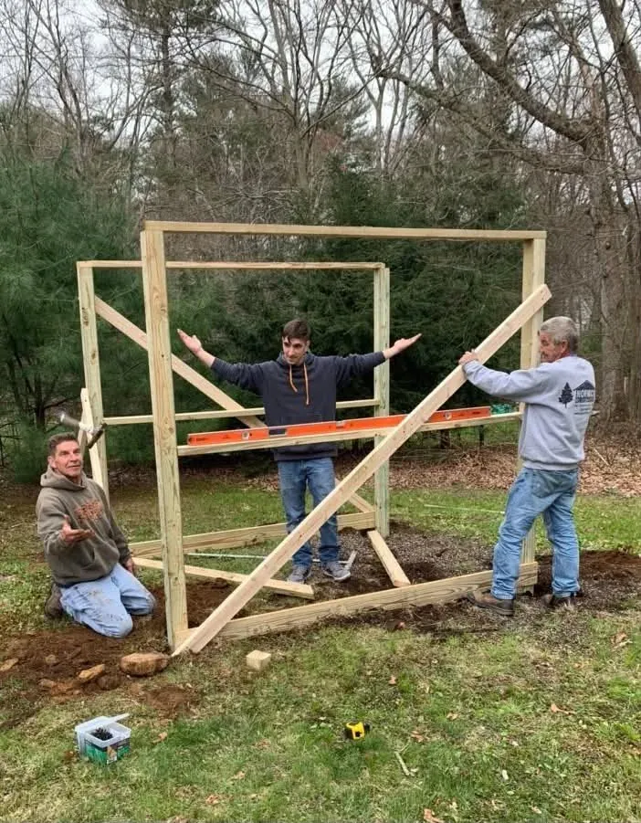 Ryan standing inside the raised wooden frame of a new enclosure with his arms outstretched, two helpers at either side